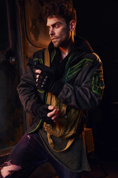 Hombre sin afeitar en chaqueta desgastada y guantes sin dedos de pie con arma de fuego en metro oscuro, post-apocalipsis - foto de stock
