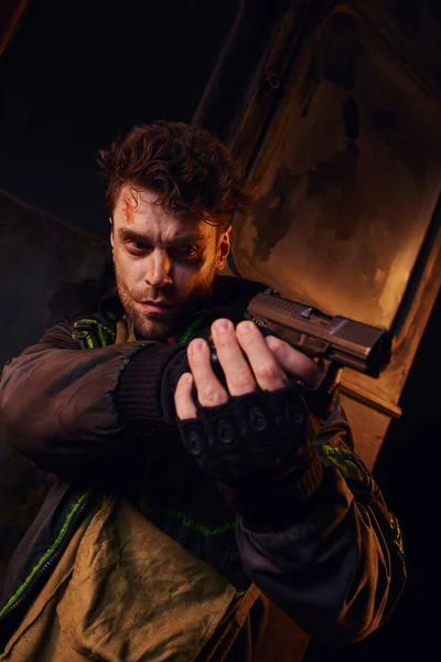 Unshaven man with scratched face aiming with gun in darkness of abandoned post-apocalyptic subway — Stock Photo