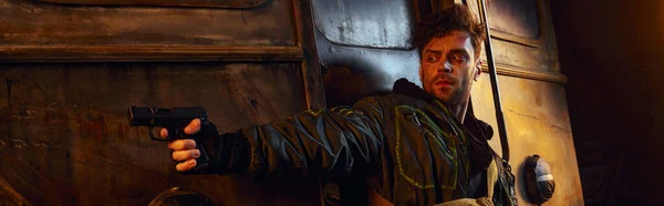 Небритый игровой персонаж в изношенной одежде, нацеленный с пистолетом в опасном постапокалиптическом метро, баннер — стоковое фото