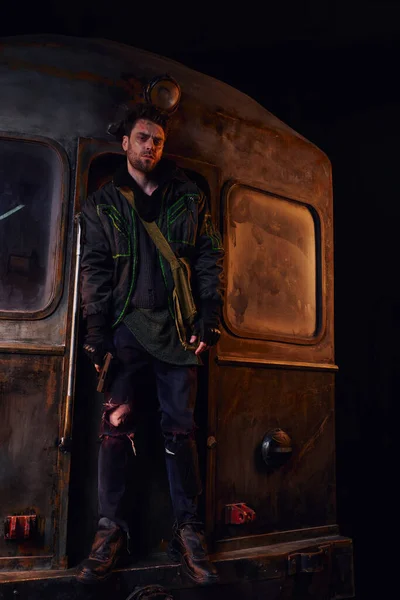 Оставшийся в живых после апокалипсиса в изношенной одежде, стоящий с пистолетом на ржавом вагоне метро в подполье — стоковое фото