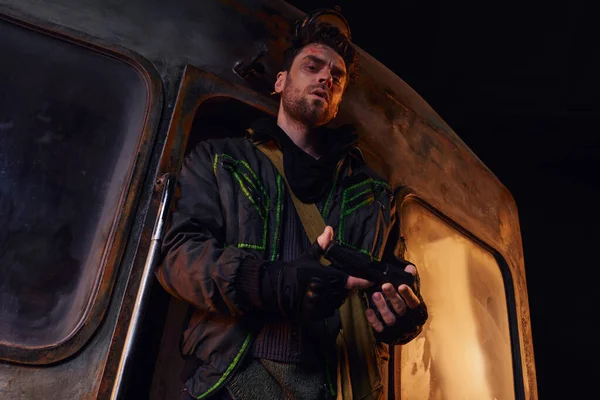 Vista de ángulo bajo del hombre sin afeitar en chaqueta desgastada mirando el arma cerca de carro oxidado en el metro abandonado - foto de stock