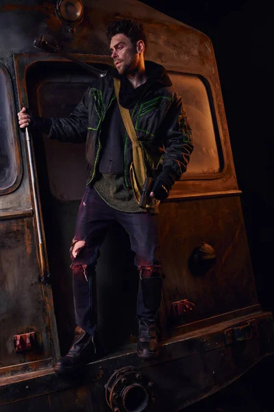 Человек в изношенной одежде, стоящий с пистолетом на ржавой карете в темном метро, постапокалиптическая антиутопия — стоковое фото