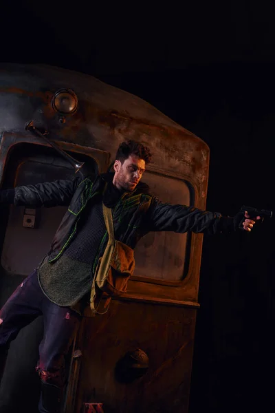 Unrasierter Mann in verschlissenem Outfit zielt mit Spaß auf rostigen Waggon in der Dunkelheit der U-Bahn nach der Katastrophe — Stockfoto