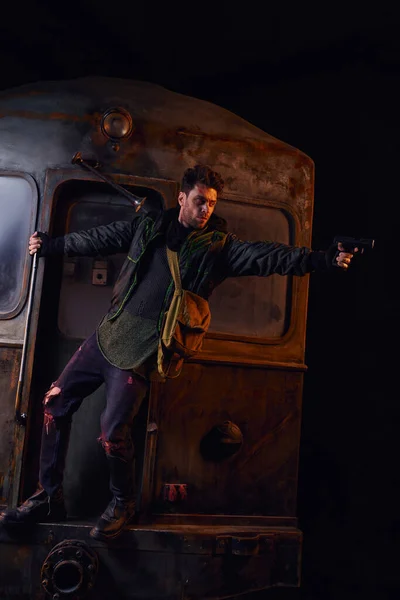 Hombre con ropa gastada apuntando con un arma a un carro oxidado en el desolado metro, peligro y supervivencia - foto de stock