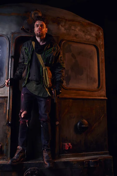 Mann in verschlissenem Outfit hält Waffe in der Nähe rostiger U-Bahn-Waggons und blickt in die Kamera, Leben nach der Katastrophe — Stockfoto