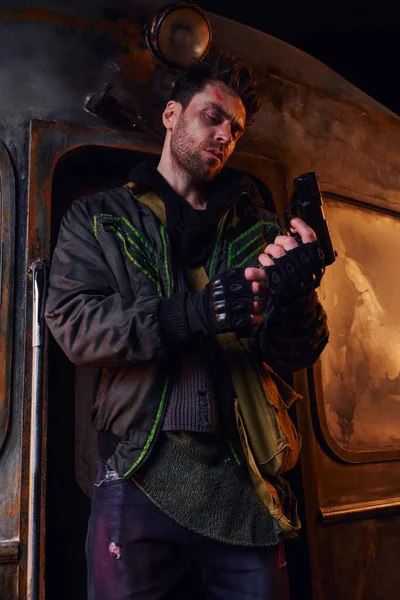 Serious man in worn jacket and fingerless gloves holding gun near rusty carriage in desolated subway — Stock Photo