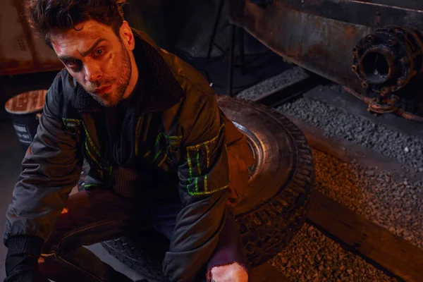 Отчаявшийся мужчина с поцарапанным небритым лицом сидит на шине в темноте постапокалиптического метро — стоковое фото