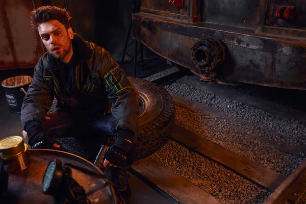 Unrasierter Mann mit Waffe sitzt auf Reifen neben U-Bahn-Waggon und Dosen mit Konserven in U-Bahn — Stockfoto