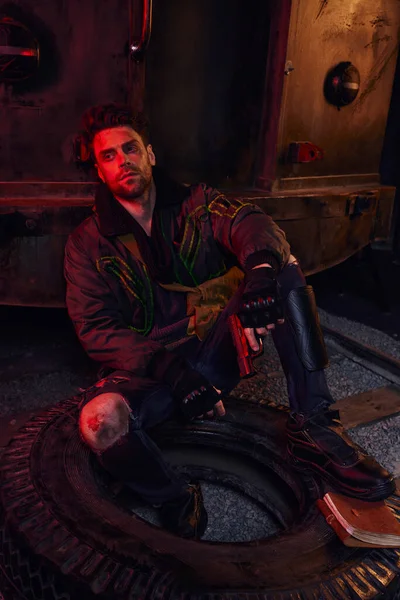 Hombre armado sin afeitar con ropa gastada sentado en el neumático cerca de los diarios en la luz roja del metro después del desastre - foto de stock
