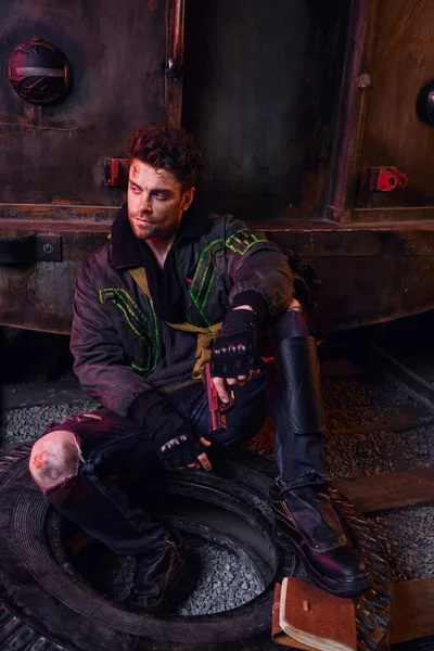 Homme non rasé avec un pistolet assis sur un pneu près de journaux intimes dans le métro sombre, concept post-apocalyptique — Photo de stock