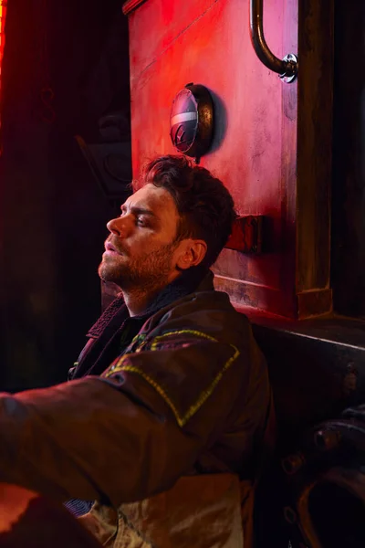 Vista lateral del hombre deprimido sin afeitar sentado cerca de un vagón oxidado en el metro, desesperación post-apocalíptica - foto de stock