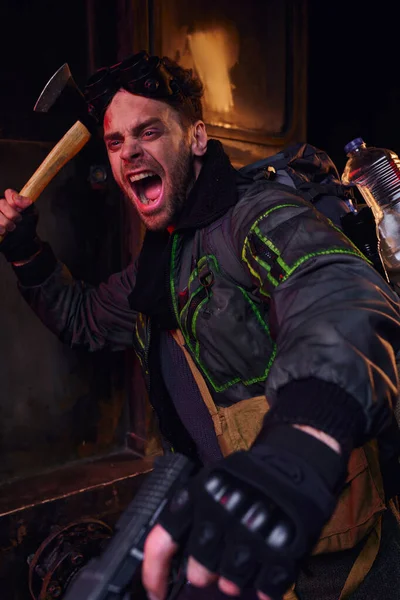 Небритый мужчина с топором и пистолетом, кричащий и защищающийся в темном постапокалиптическом метро — стоковое фото