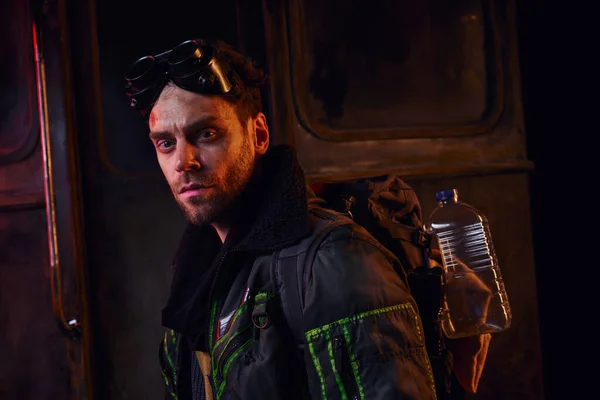 Man with military goggles, backpack and water bottle looking at camera in post-apocalyptic subway — Stock Photo