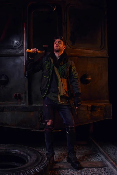 Людина в одягненому брудному одязі, що стоїть з сокирою біля іржавої каретки метро, після катастрофи виживання — стокове фото