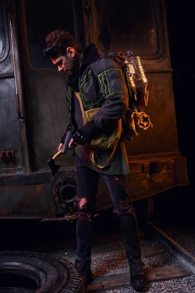 Mann mit Waffe und Axt bewaffnet steht mit Rucksack und Wasserflasche in dunkler verlassener U-Bahn — Stockfoto