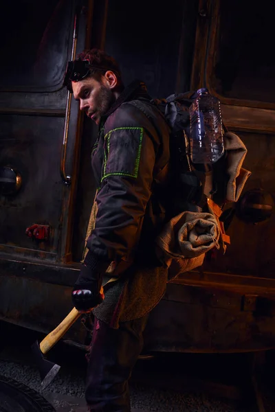 Seitenansicht von grimmigem Mann mit Rucksack und Wasserflasche, der mit Axt in gefährlicher verlassener U-Bahn steht — Stockfoto