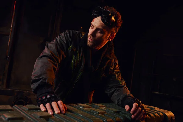 Небритый человек с гуглами, смотрящий в сторону ящика с оружием в темноте постапокалиптического подполья — стоковое фото