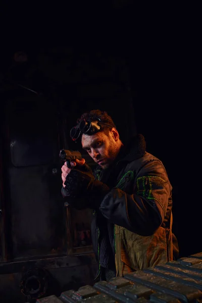 Homme avec des lunettes militaires regardant loin et visant avec arme à feu à la lumière rouge du métro post-apocalyptique — Photo de stock