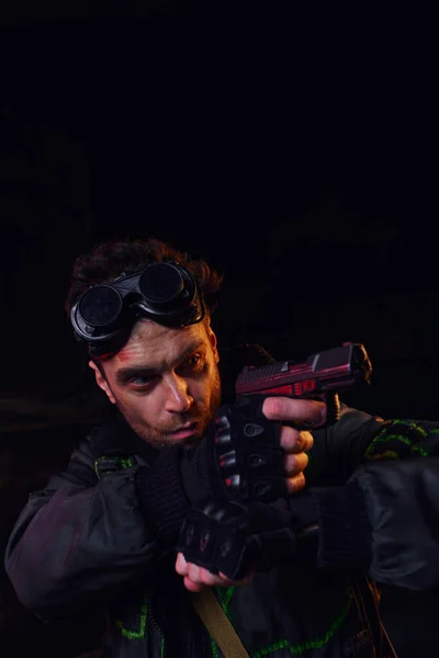 Концентрированный человек с военными гуглями, нацеленный с оружием в темноте постапокалиптического подполья — стоковое фото