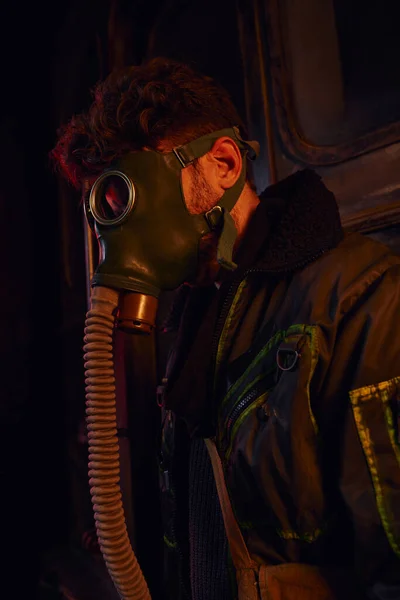 Hombre con máscara y chaqueta desgastada en luz roja de túnel subterráneo oscuro, supervivencia post-apocalíptica - foto de stock