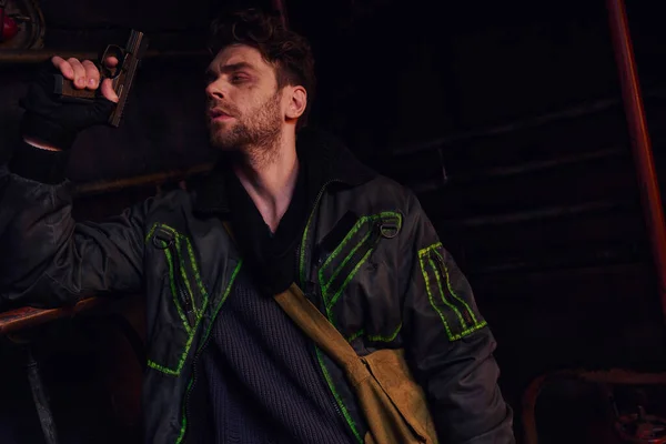 Небритый мужчина в изношенной одежде, держащий пистолет и смотрящий в опасную темноту заброшенного метро — стоковое фото