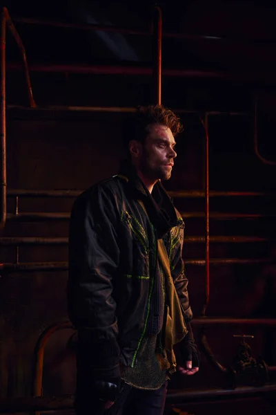 Unshaven man in black jacket looking away near rusty pipes in post-disaster underground tunnel — Stock Photo