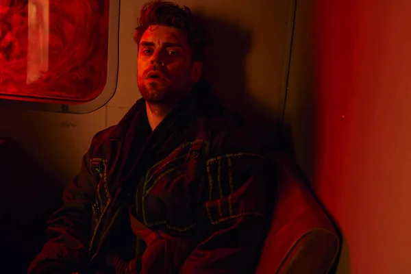 Измученный человек, сидящий в красном свете грязной кареты постапокалиптического метро, игровой персонаж — стоковое фото