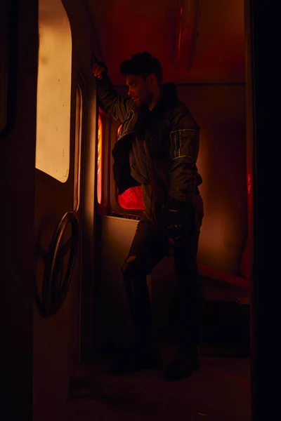 Uomo con pistola che guarda attraverso il finestrino della carrozza abbandonata nella metropolitana post-disastro con luce rossa — Foto stock