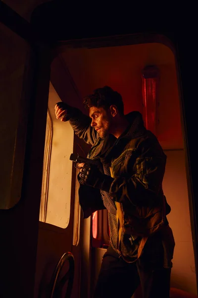 Человек с пистолетом стоит у окна заброшенной кареты в постапокалиптическом метро с красным светом — стоковое фото
