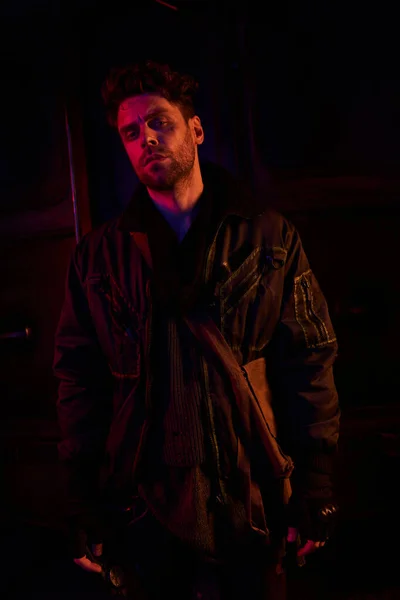 Sombrío hombre de chaqueta negra desgastada mirando a la cámara en el oscuro metro post-apocalíptico con luz roja - foto de stock