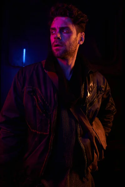 Tense man in black jacket looking away in darkness of abandoned underground tunnel with neon light — Stock Photo