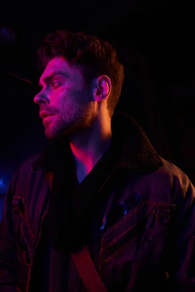 Despaired unshaven man in jacket looking away in neon light of dark post-apocalyptic underground — Stock Photo