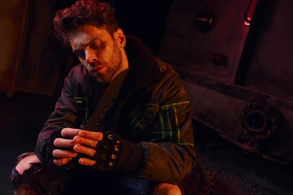 Небритый мужчина в перчатках без пальцев держит военный нож в подземелье после катастрофы с красным светом — стоковое фото