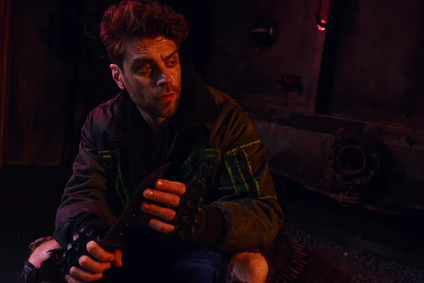 Uomo premuroso in guanti senza dita seduto con coltello militare nel sottosuolo scuro con luce rossa — Foto stock