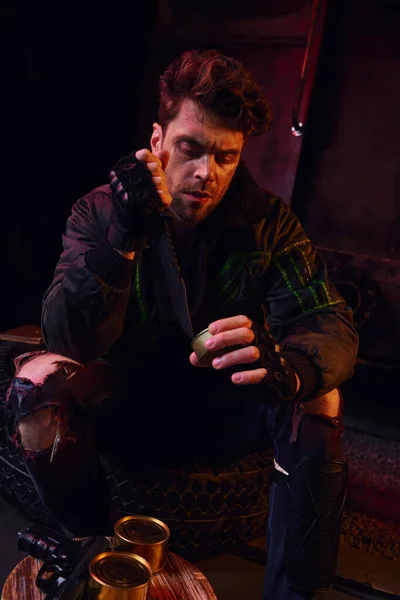 Голодный человек в изношенном наряде открывает консервы с военным ножом в темном подполье после катастрофы — стоковое фото