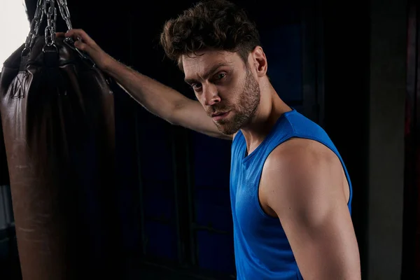 Self-assured sportive man in blue tank top looking at camera near punching bag on night street — Stock Photo