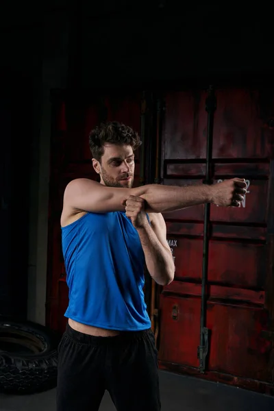 Sportive unshaven man in blue tank top stretching arm in city at night, street sport — Stock Photo
