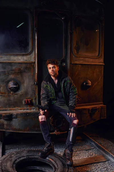 Hombre sin afeitar en traje desgastado sentado con el arma cerca de carro oxidado y neumático en el metro después del desastre - foto de stock