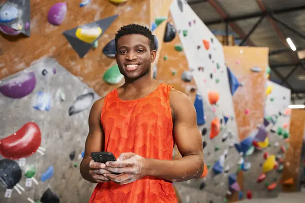 Jolly african american man in orange shirt holding mobile phone and looking at camera, rock climbing — Stock Photo