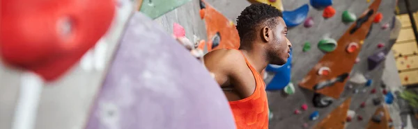 Handsome african american man in orange shirt next to rock climbing wall looking away, banner — Stock Photo