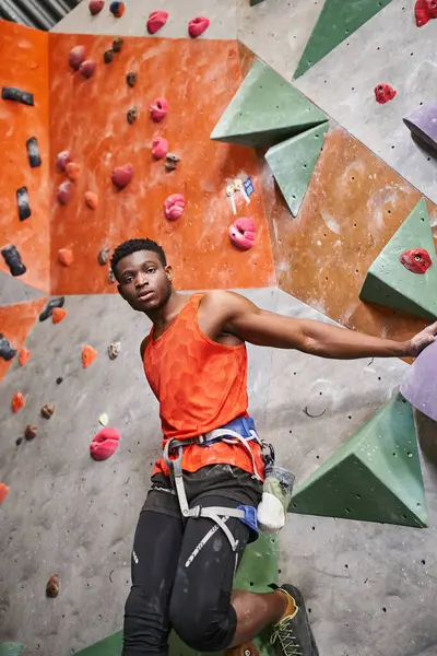 Handsome african american man with alpine harness in orange shirt posing next to rock climbing wall — Stock Photo