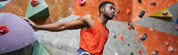 Handsome african american man with alpine harness posing near climbing wall and looking away, banner — Stock Photo