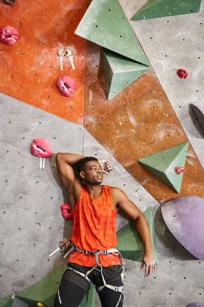 Tiro vertical del hombre afroamericano cerca de la pared de escalada con la mano detrás de la cabeza, bouldering - foto de stock