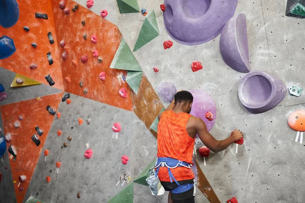 Forte afroamericano in camicia arancione arrampicata su parete rocciosa con imbracatura alpina, bouldering — Foto stock