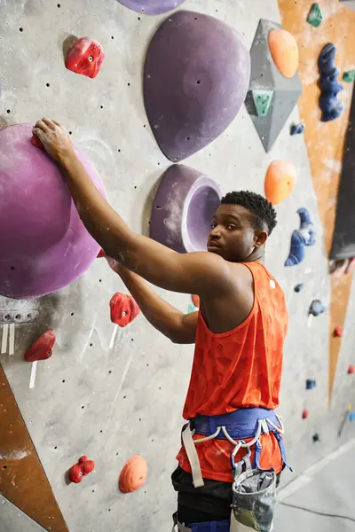 Colpo verticale di uomo afroamericano con imbracatura alpina in posa su parete da arrampicata, sportivo — Foto stock