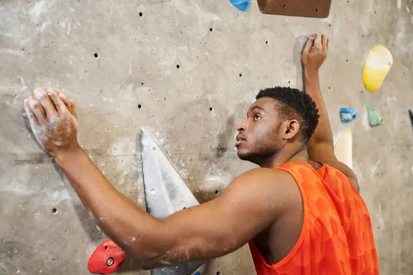 Hombre americano africano musculoso guapo en camisa anaranjada que sube encima de la pared de roca, concepto de bouldering - foto de stock