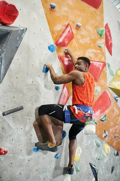 Vertical shot of young african american man in orange shirt climbing up wall and looking ta camera — Stock Photo