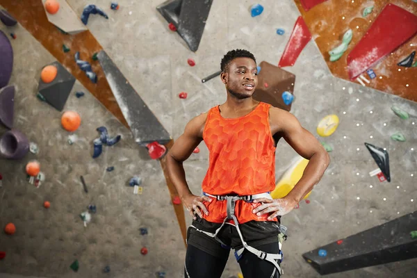 Atletico afroamericano in camicia arancione con imbracatura alpina in posa con le mani sui fianchi — Foto stock