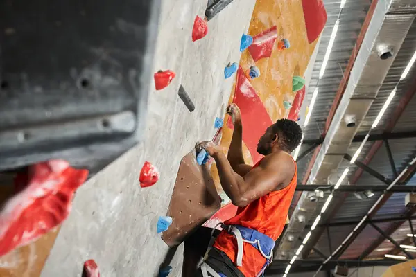 Handsome african american man in orange shirt with alpine harness climbing up bouldering wall — Stock Photo
