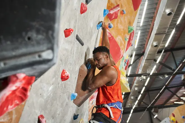 Handsome young african american male model with alpine harness climbing up bouldering wall — Stock Photo